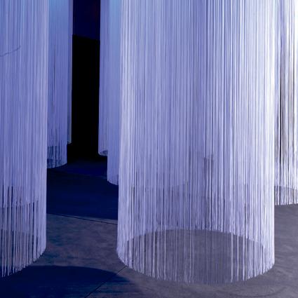 Hvide trådgardiner, der hænger i cirkler i et rum med blåt lys. Der er flere gardincirkler og tilsammen danner de nærmest en kunstinstallation.