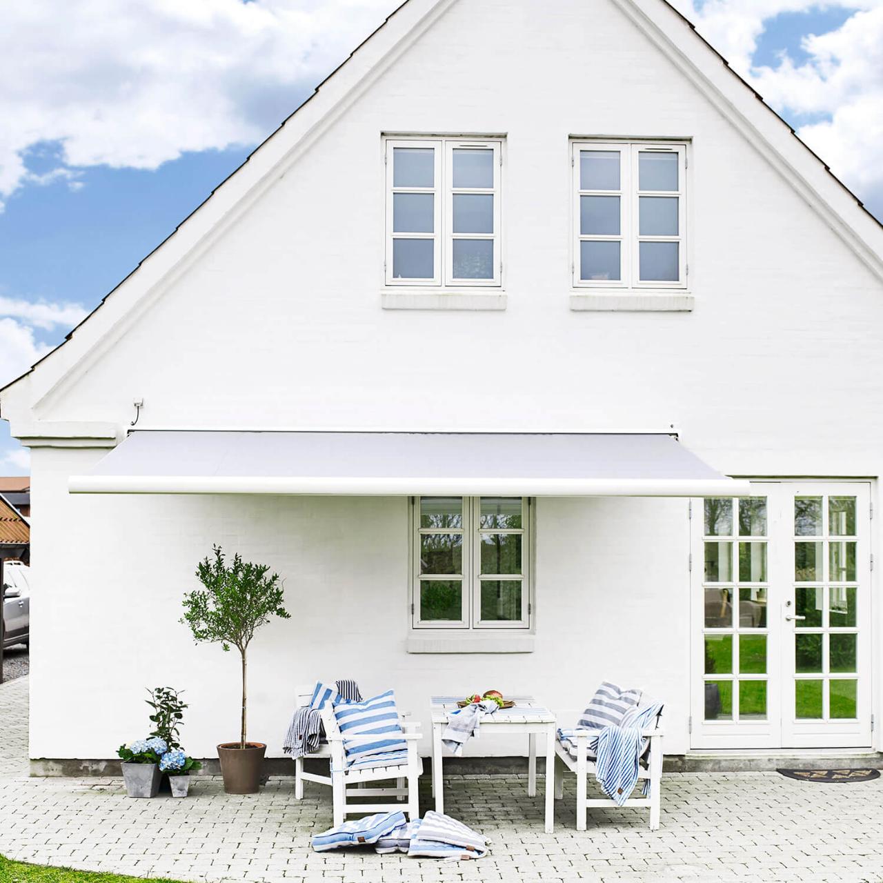 Hvidt hus med hvide vinduer og en markise hen over terrassen, hvor der står et hvidt havebord med træstole.